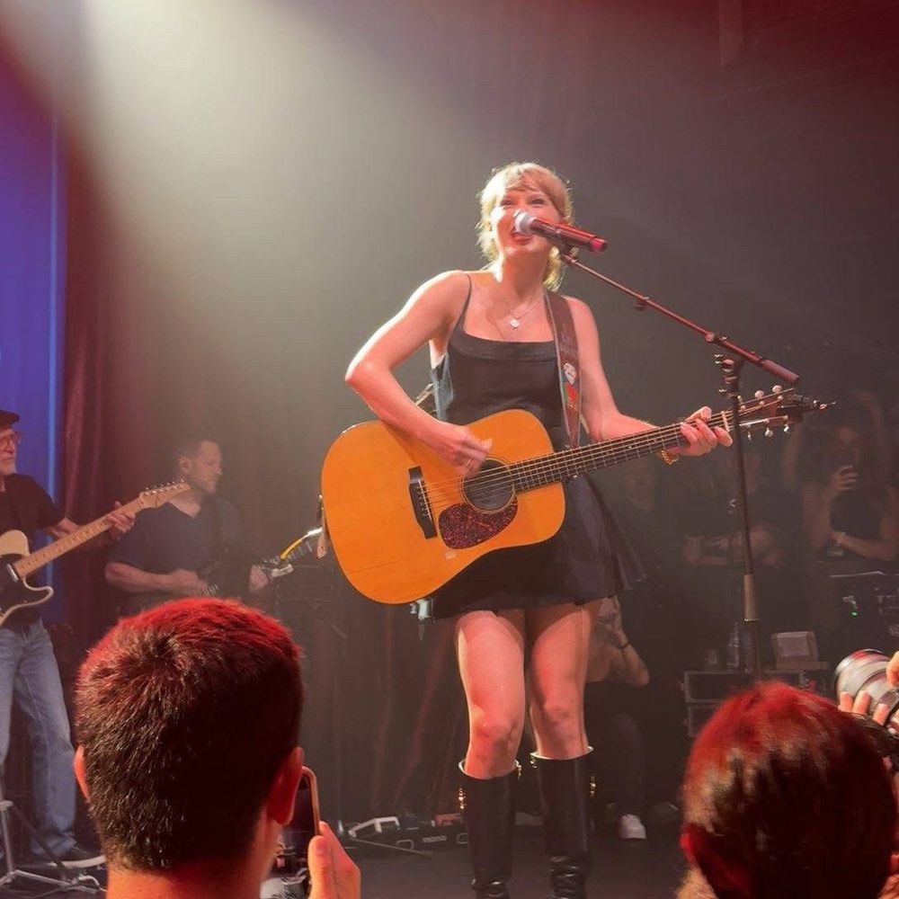 Current day Taylor Swift holding a brown guitar singing into a stand up microphone with tied up blonde hair, a black thigh length dress and standing in front of an audience with musicians holding guitars in the background  