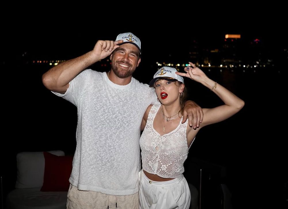 Taylor Swift and travis kelce wearing saying captain holding them, both are wearing white tops and white/biege bottoms. It is nightime and there are blurred lights behind them 