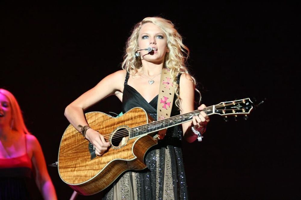 Young taylor swift holding a brown guitar wearing a black and gold dress with blonde curly hair and a microphone attached to her ear 
