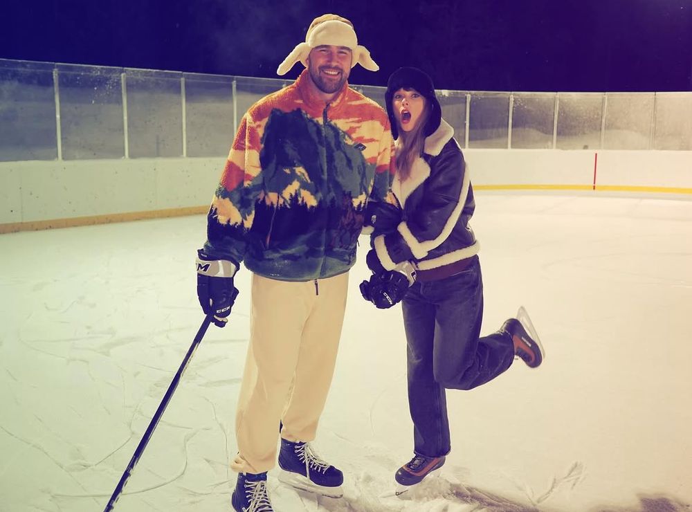 Travis kelce and taylor swift smiling in snow, he is wearing a multicoloured fleece and white jogging bottoms and taylor is wearing a black and white leather jacket and jeans 