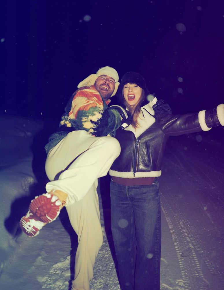 Travis kelce and taylor swift smiling in snow, he is wearing a multicoloured fleece and white jogging bottoms and taylor is wearing a black and white leather jacket and jeans 