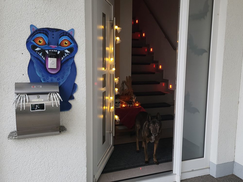 Offene Haustür, mit  Gespenster-Lichterkette dekoriert. Innen steht ein Tisch mit Knochen-Deko, zwei Kerzen und einer großen Schale mit Geschenktüten für die Kinder. Der Briefkasten und die Klingel neben der Haustür sind mit dem selbstgebastelten Derpy dekoriert.