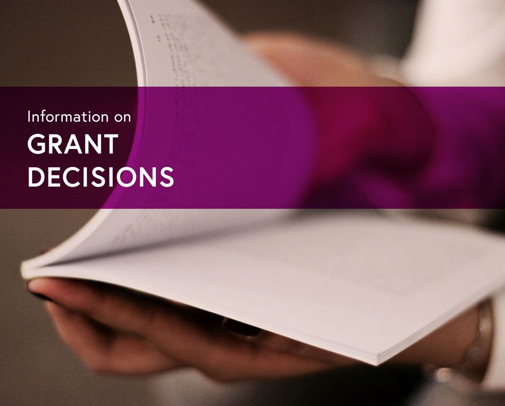 A person's hands hold a paper booklet, open for reading. The background is blurry, and you cannot see any faces. The text on the photo reads Information on Grant Decisions.
