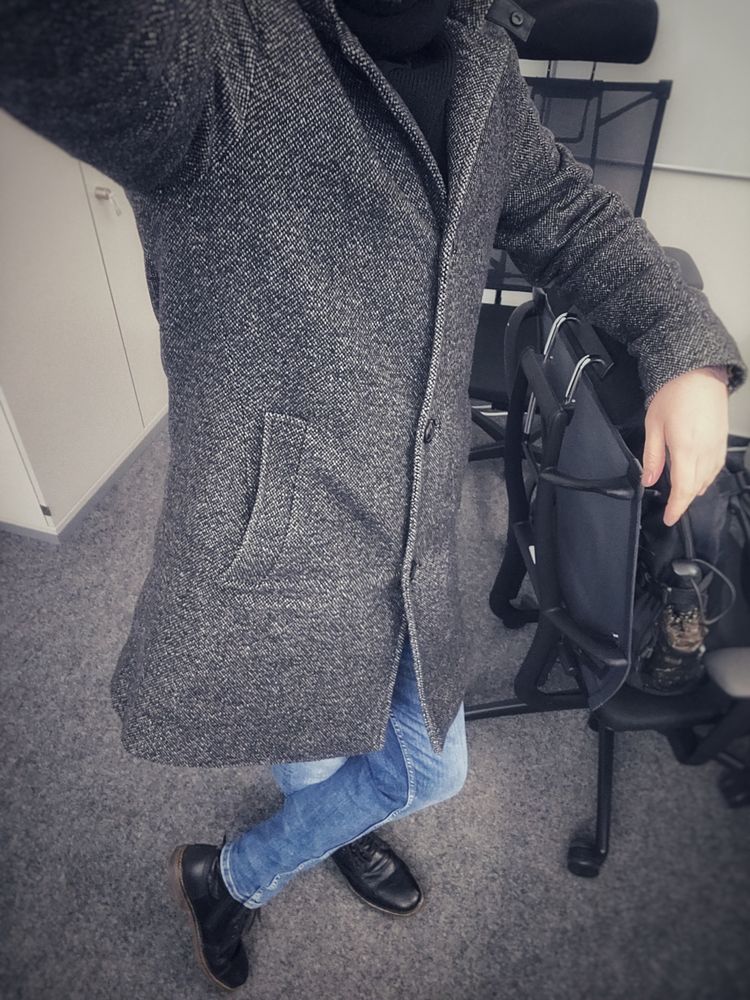 Ich stehe an einen Schreibtischstuhl gelehnt im Büro.
Ich trage schwarze Schuhe, eine blaue Jeans und einen grauen Mantel.