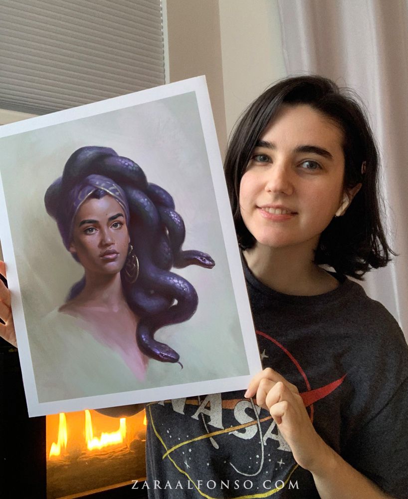 Photograph of the artist Zara Alfonso holding a print of her painting "Medusa Portrait".  A portrait of a young Medusa who looks peacefully towards the viewer, her hair is flowing mass of thick iridescent dark indigo snakes tied up in a head wrap.