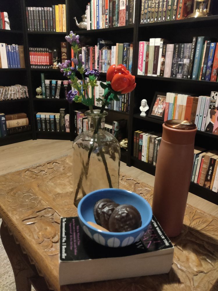 Bilde av en liten, blå skål med Jaffakjeks i, en bok, en vase med Lego-blomster og en drikkeflaske på et brunt bord. I bakgrunnen sees bokhyller.