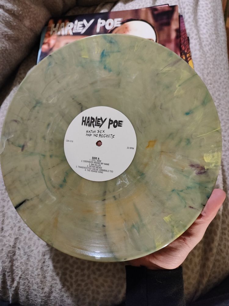 Me holding a light brown-ish green vinyl with a bunch of random multi-colored splashes. The vinyl is labelled Satan, Sex, and No Regrets by Harley Poe, side A.