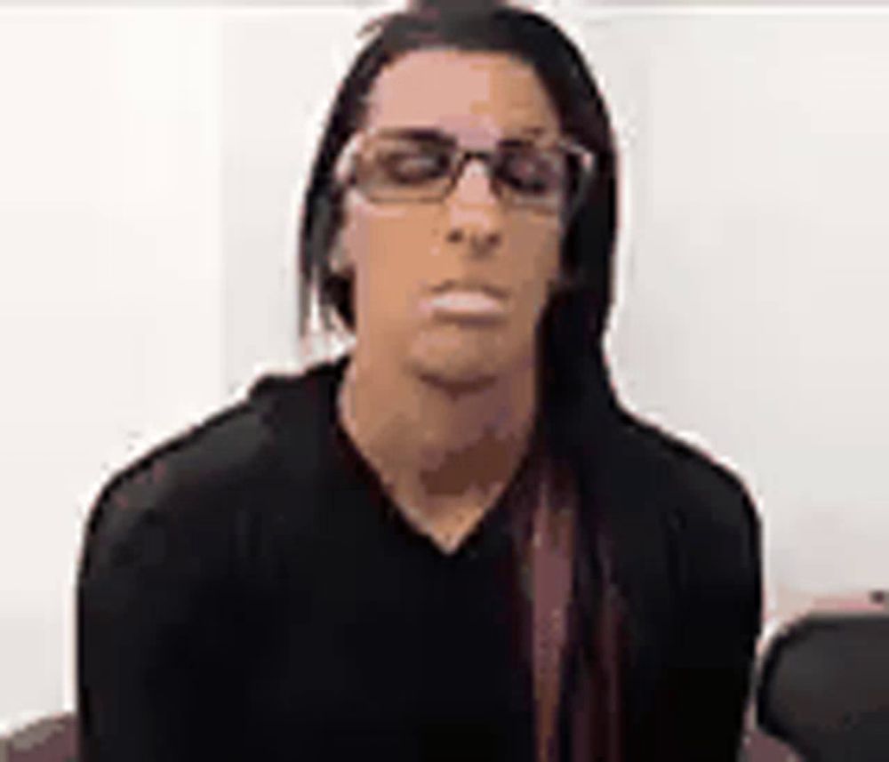 a man with long hair and glasses is wearing a black shirt and making a funny face .