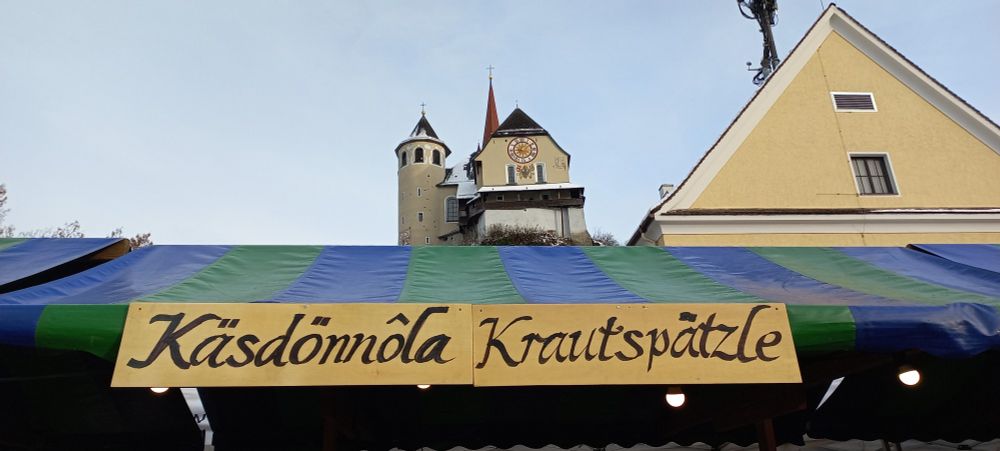 Foto von Verkaufsstand Adventmarkt der Pfadfinder Rankweil mit dem Titel: Käsdönnala, Krautspätzle, im Hintergrund ist die Basilika von Rankweil zu sehen.
