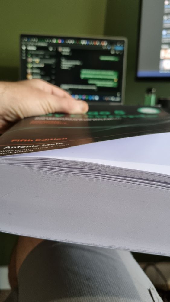 A book side on showing that it is a very thick book. In the background is a blurry laptop screen and beneath the desk the book is placed on is a man's leg.