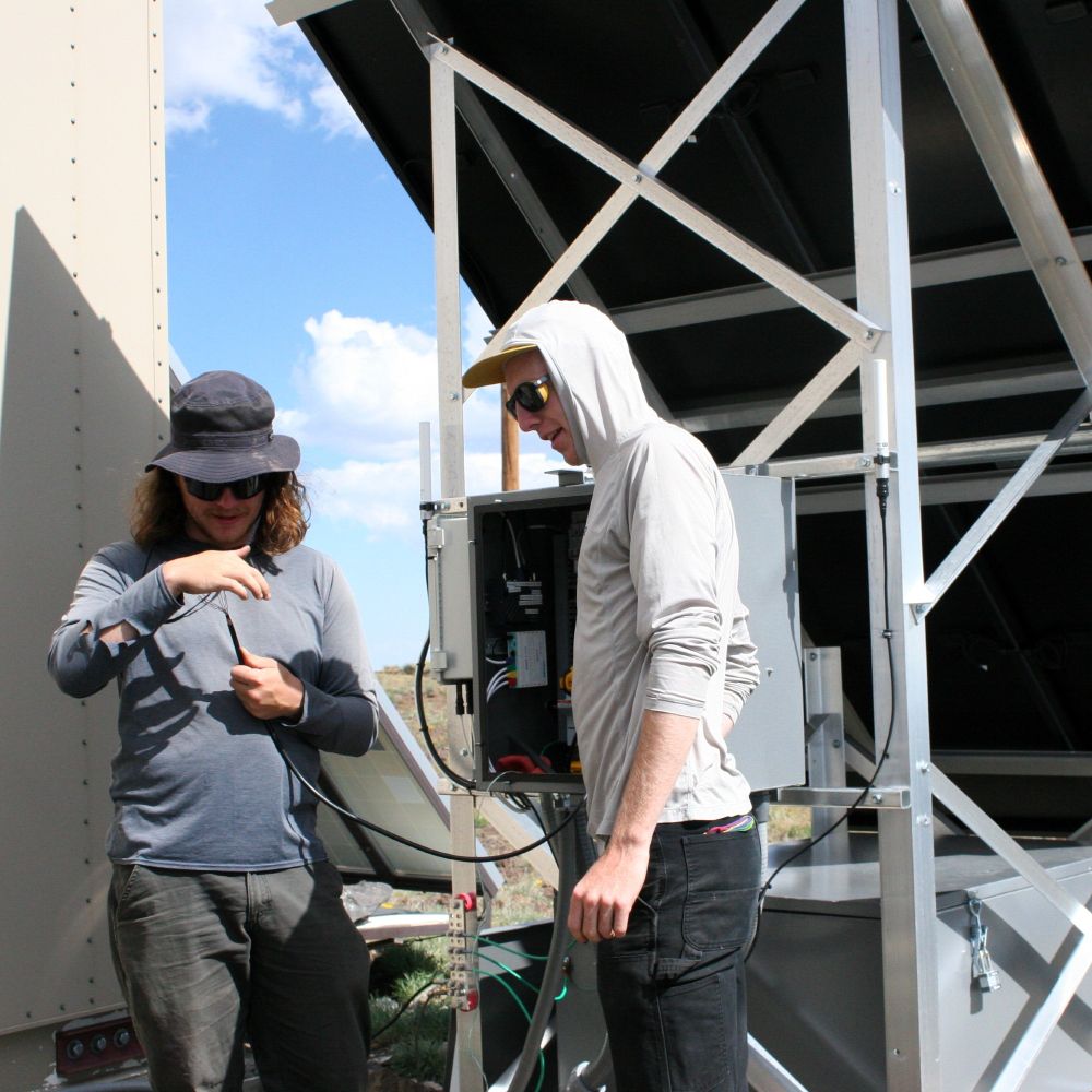 Two young men repair a wildfire camera on a desert mountaintop.