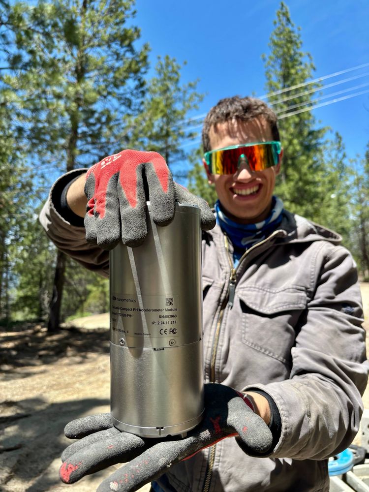 A young man holds a seismic sensor while posing for a photo in a forest.