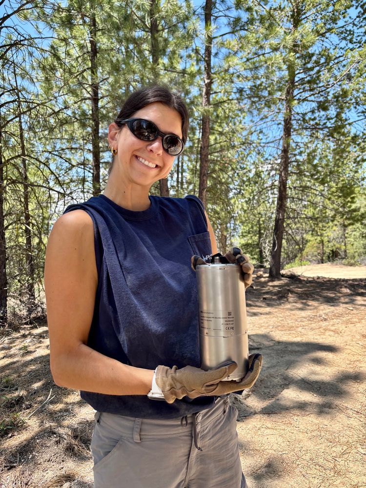 A young woman holds a seismic sensor while posing for a photo in a forest.