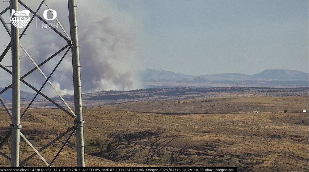 A tall column of white smoke rises in the distance behind a grassy landscape and a communications tower, captured by the Shaniko camera during daylight.