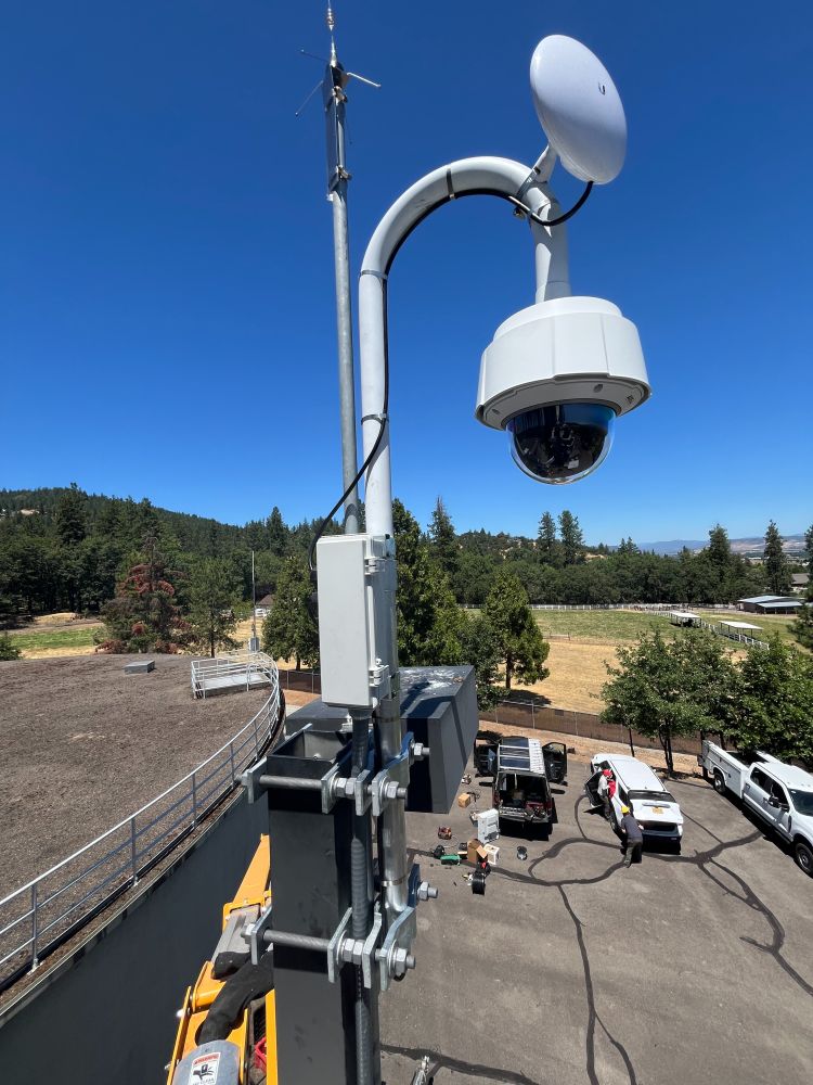 A wildfire detection camera is on a building with views of forests and hills in the background.