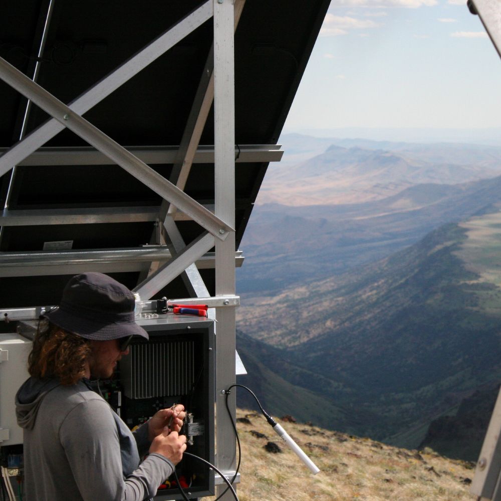 A young man repairs a wildfire camera on a desert mountaintop.