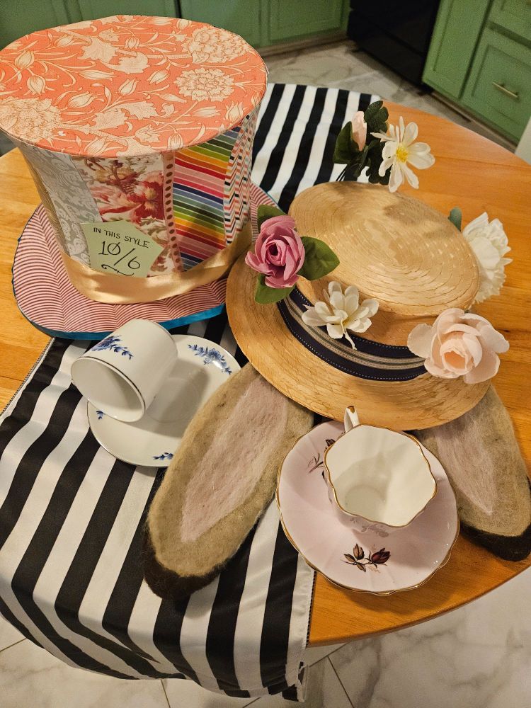 A colorful and many-patterned tophat with a "In This Style 10/6" tag and a straw hat with flowers in the band and rabbit ears sticking out of the bottom sit on a table with a black-and-white striped runner. Two teacups sit on saucers next to the hats.
