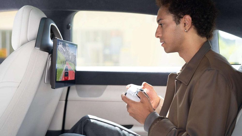 A young man in the back of a car, playing a game with a DualSense controller from a screen integrated into the back of the front seat in front of him