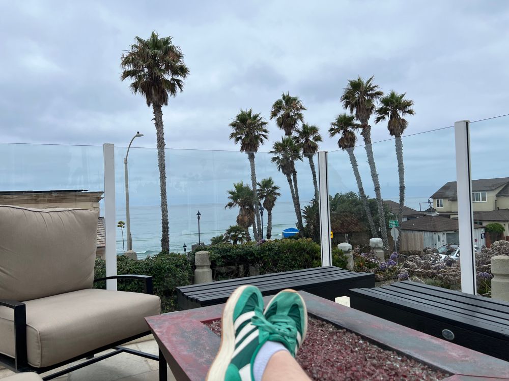 my feet resting on a fire pit with some tasteful sock showing. in the distance is some palm trees and the ocean 