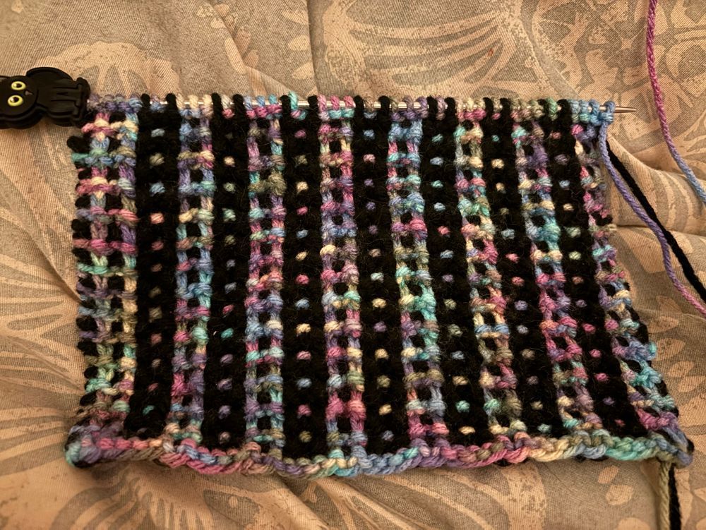 A rectangular knitting project worked in mosaic knitting. One color is black and the other yarn is a variegated pink, blue, purple, and yellow.