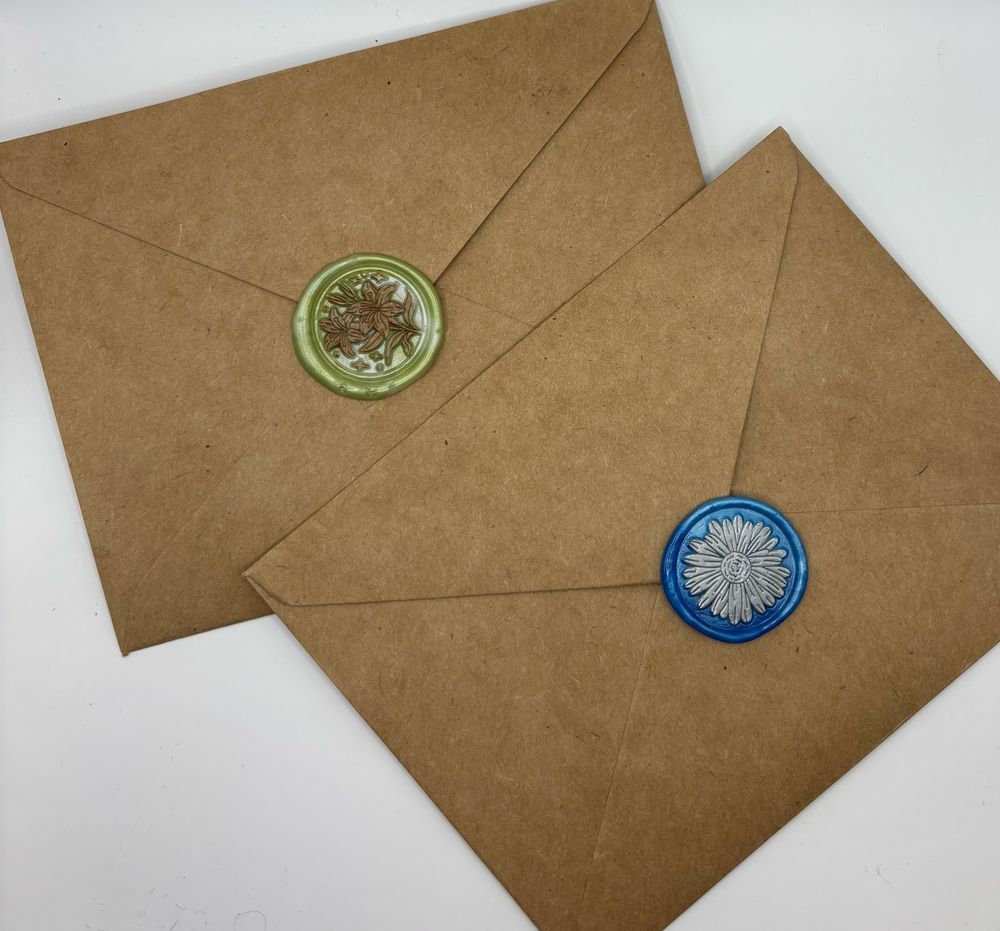 Two brown paper envelopes with wax seals. The first envelope has a light green wax seal with golden lilies. The second wax seal is a light blue with a silver daisy.