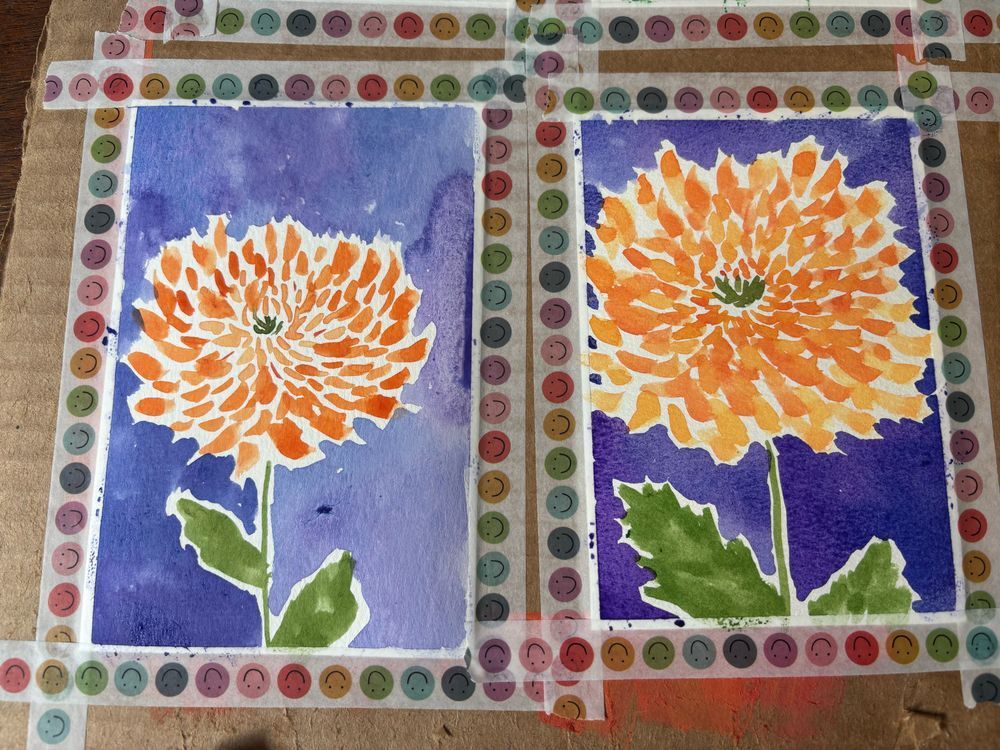 Two pieces of watercolor paper taped down to a board with washi tape. Each piece of paper has an orange chrysanthemum painted on it with a green stem and leaves, and a blue and purple background.
