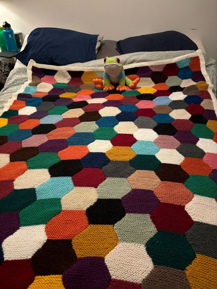 Knit blanket made of sewn-together hexagons in different colors. The blanket is edged in a white garter stitch border. The blanket is lying on a bed, and a stuffed frog sits on top.