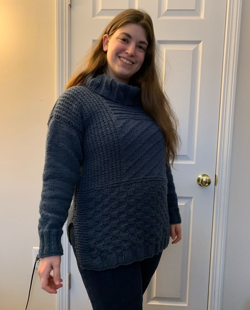 Woman standing and wearing a blue turtleneck knit sweater. The sweater has multiple patterns of knit and purl stitches.