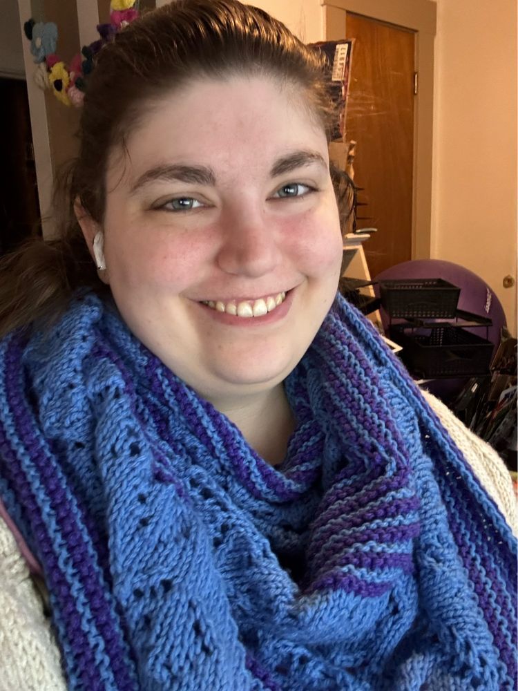 A woman wearing a blue and purple knit shawl. The shawl alternates between garter stripes in blue and purple and a solid blue lace section.