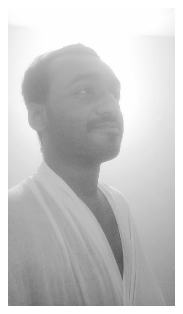 Black-and-white photo of a person in a soft, glowing light. The individual is wearing a light, draped garment and looking slightly upward with a calm, thoughtful expression. The background is minimal and bright, creating a serene and ethereal atmosphere.