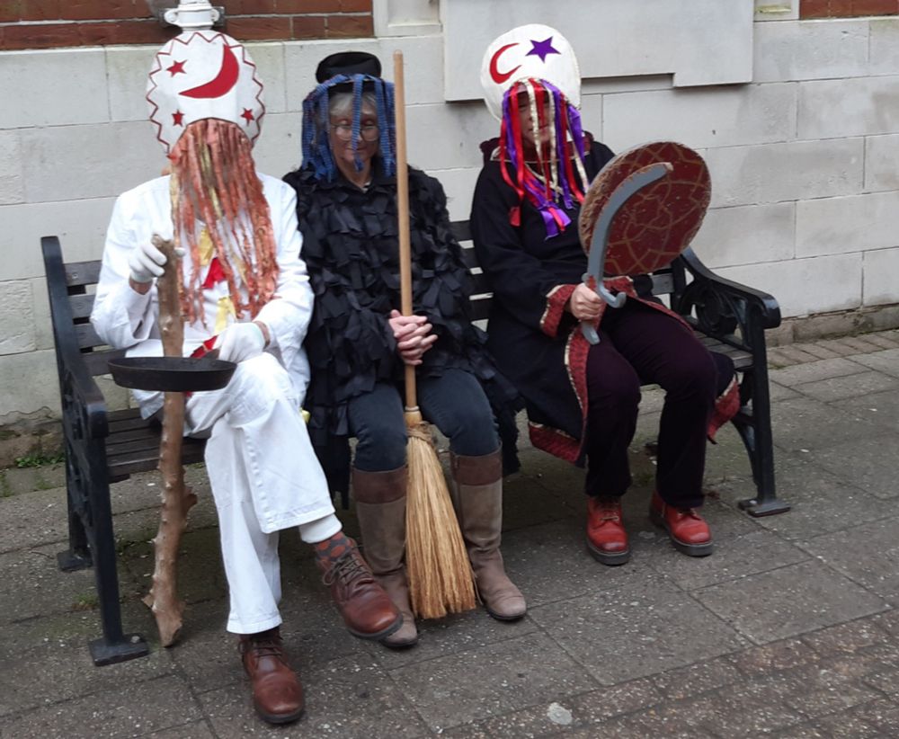 Three odd characters sit on a bench. At left, all in white with a club in his hand and wearing a comical hat decorated with a moon and stars, and with ribbons obscuring his face is Beelezebub. In the middle dressed in black with a crumpled top hat, also with ribbons obscuring their face is the Introducer. Finally on the right is the Turkish Knight, with a navy woollen robe over trousers and holding a scimitar. All from the Dorset Mummers play...