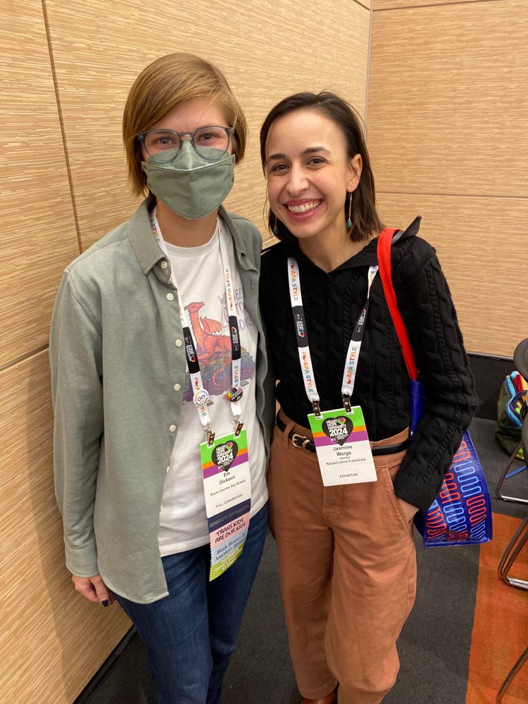 A photo of me, a white person with short sandy hair and pale green glasses, wearing a pale green face mask and matching button-down shirt worn open over a white We Need LGBTQ+ Books dragon tee, standing with acclaimed author Jasmine Warga, whose most recent middle grade book is entitled A Strange Thing Happened in Cherry Hal