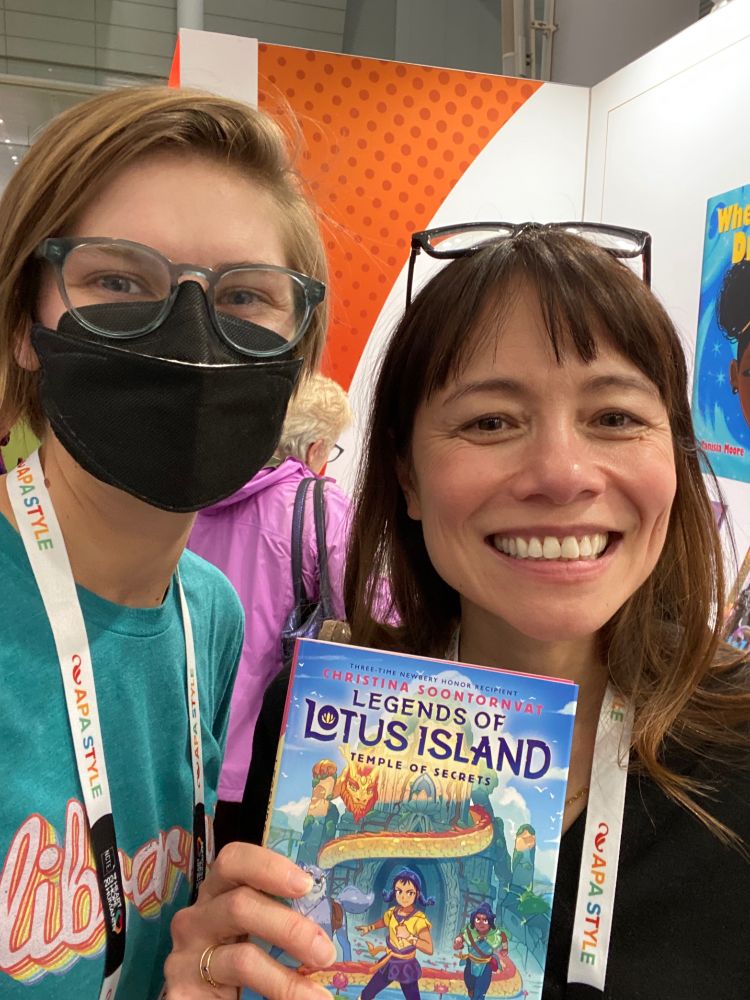 A selfie of me, a white person with short sandy hair and pale green glasses, wearing a black face mask and a teal shirt that reads “librarian” in rainbow letters, and acclaimed author Christina Soontornvat as she holds up the most recent book in her middle grade Legends of Lotus Island series - Book 4, Temple of Secrets