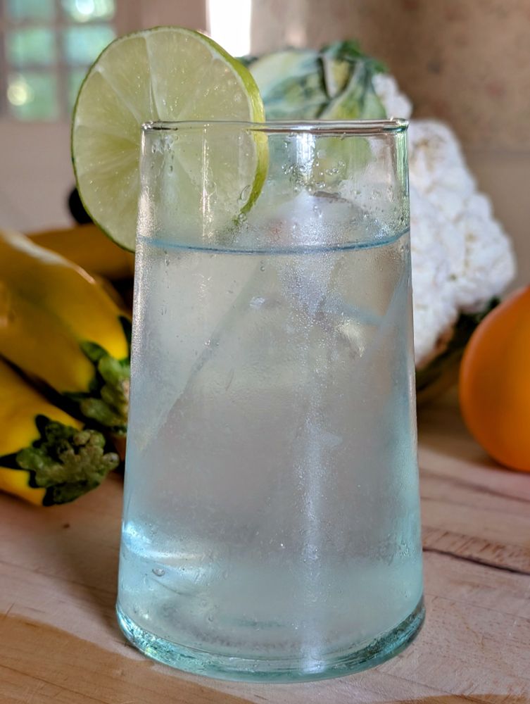 gin and tonic in a glass with a slice of lime, on a wooden cutting board with vegetables in the background 