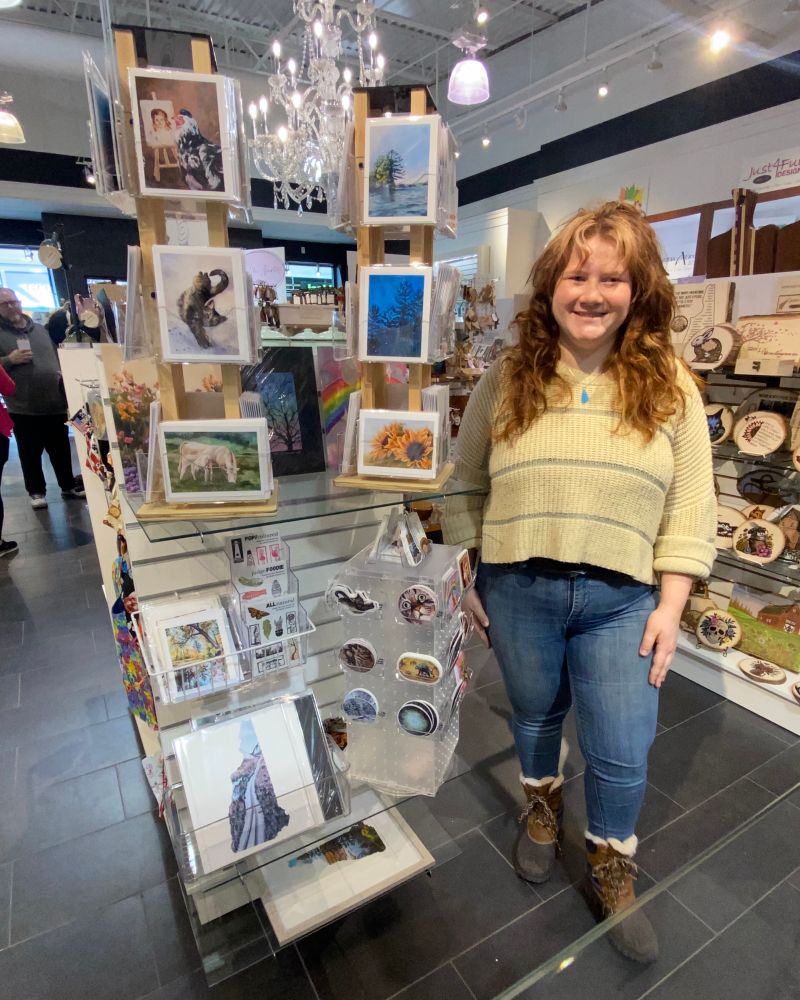 A smiling artist stands beside a retail display of her greeting cards, stickers, and art prints. 