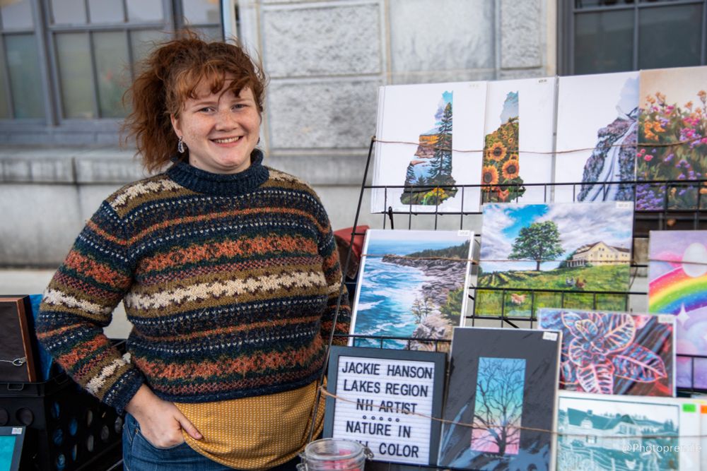 A smiling redheaded woman in a patterned sweater stands next to a display of colorful art prints.