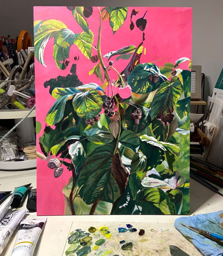 An unfinished painting of a green leafy plant over a flat, hot pink background. Details from the artist’s desk are in view, including paint tubes, a dirty palette, and brushes. 