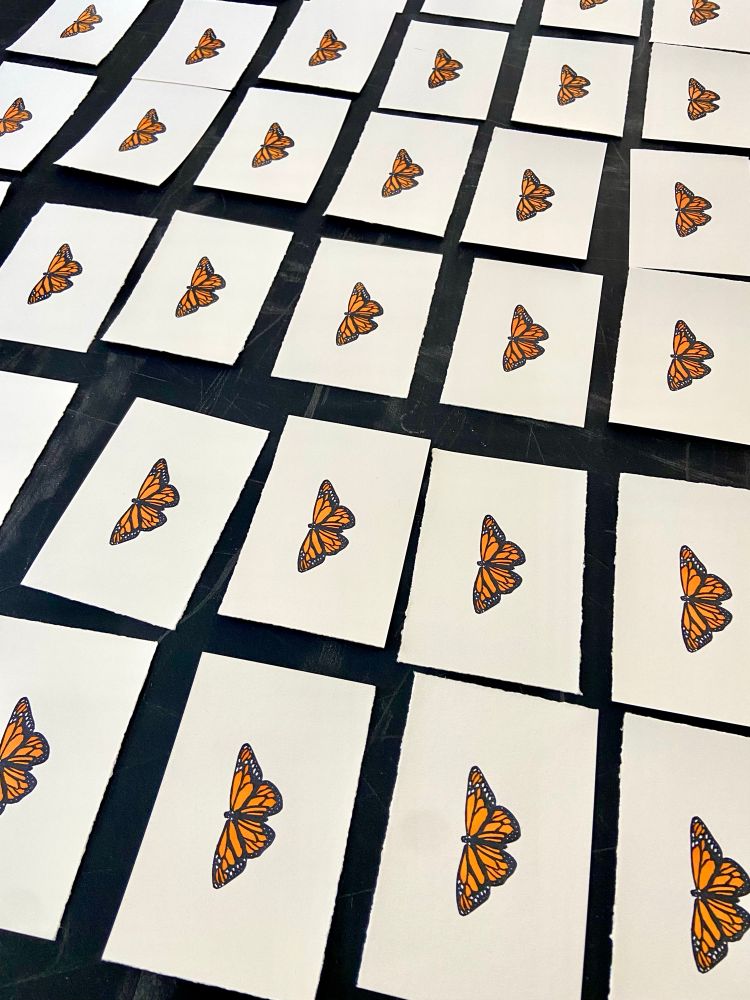 An array of small rectangles of paper, each with a hand-printed monarch butterfly, colored in orange, at the center. 