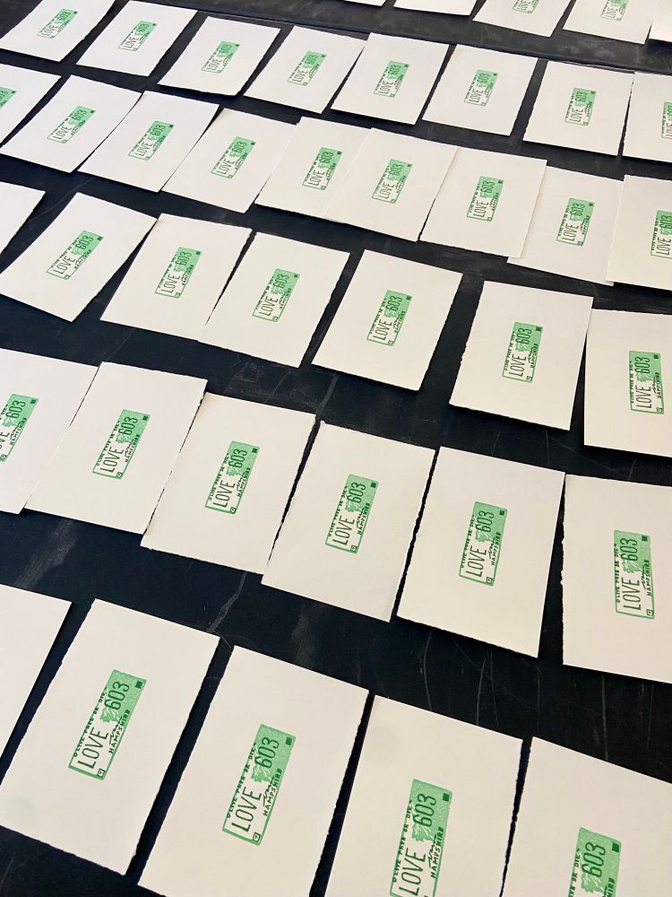 An array of rectangles of paper, each hand-printed with a stylized version of the New Hampshire license plate reading “LOVE 603”. 