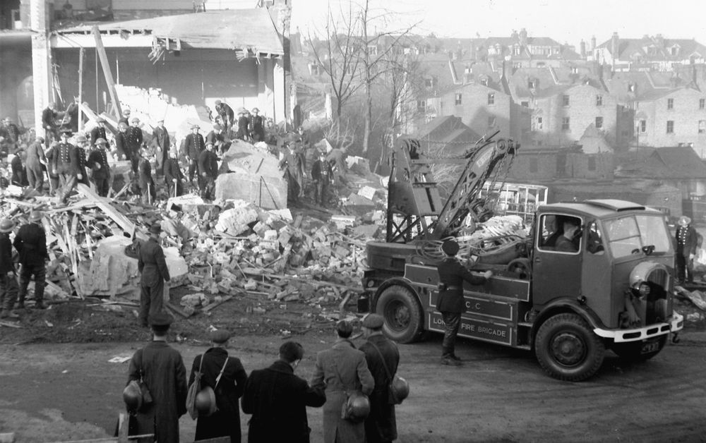 Firemen and Rescue Squad workers sift through the ruins of Invicta Road School whilst a group of civilians watch the work.