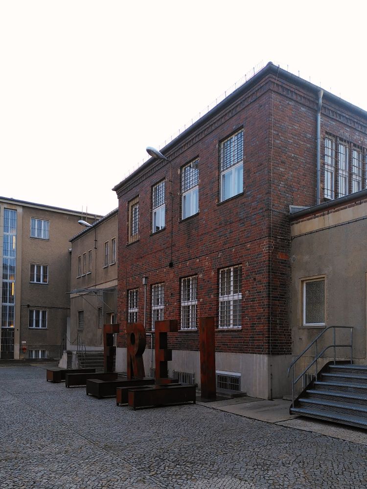 Blick von einem gepflasterten Hof auf ein Backsteingebäude mit vergitterten Fenstern. Davor in rostigem Metall große Buchstaben stehend FREI liegend HEIT