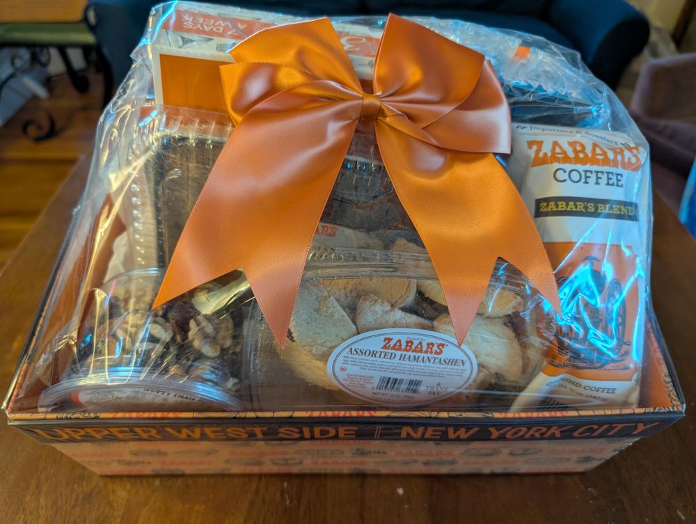 Gift box from Zabar's. You can barely make out "Upper West Side New York City" on the front of the box. It's wrapped in plastic, has a big orange bow on top, and has visible delicacies including nut mix, hamentashen, coffee, and other things you can't make out.
