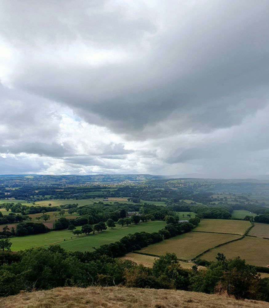 Below dramatic dark clouds filtering sunshine over patchwork of fields and trees. 