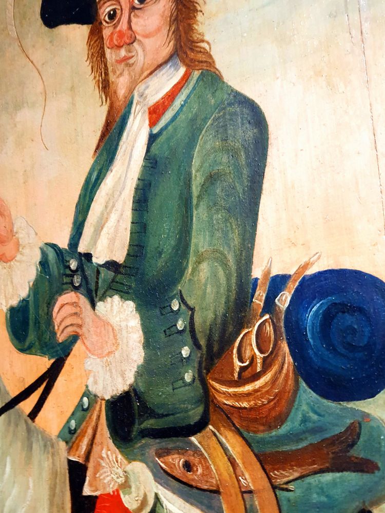 Colourful male character, red nose, riding goat, wearing hat, deep cuffed green jacket & white shirt with frilly cuffs. Red herring and leek (partly visible) strapped to saddle; and blue bed roll. From Inn Sign.