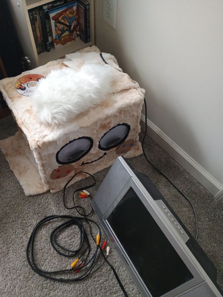 A mostly finished goob head with freshly-sewn cheeks looks at an old TV with a bundle of rca cables nearby.