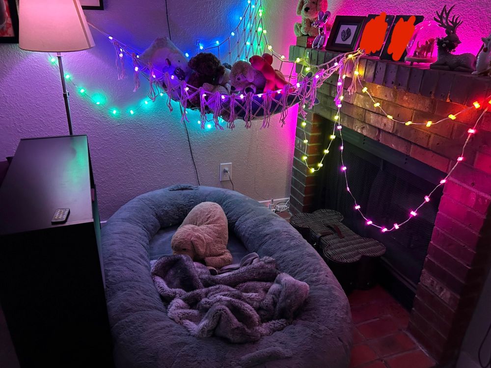 a picture of a dog bed in front of a fireplace with a plushie hammock, a bookshelf, and string lights, there’s a blanket and puppy plushie in the dog bed