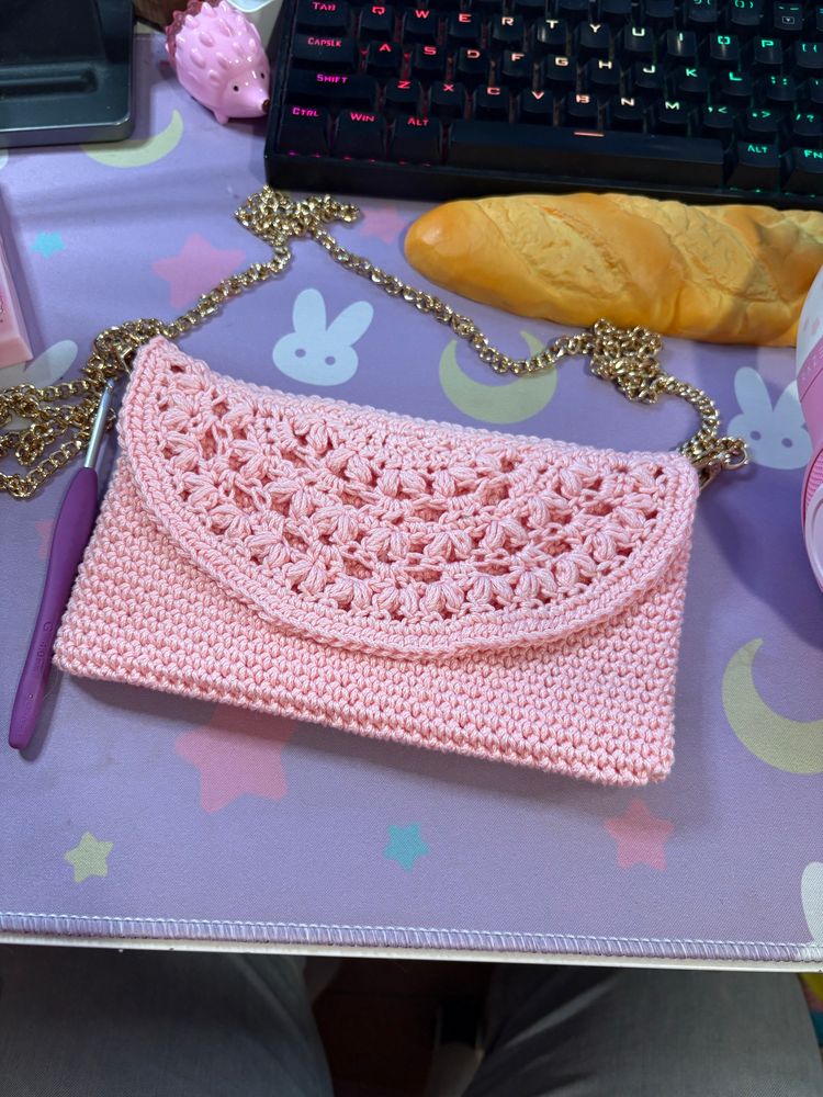 top down view of a desk, there is a with a light pink crocheted clutch purse with a gold chain, around it there are a purple-handled crochet hook, pink hedgehog-shaped pencil sharpener rainbow rbg keyboard, foam baguette keyboard rest 