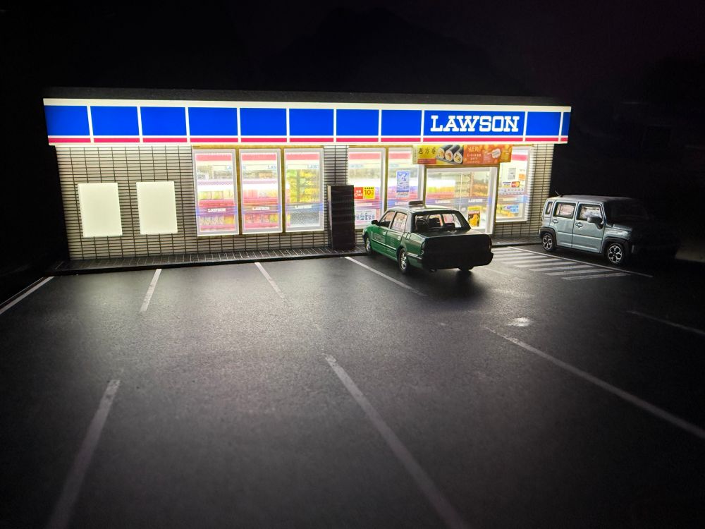 A small illuminated model of a Lawson convenience store, like one would see in Asia. It’s at 1:64 scale, and parked outside is a Toyota Crown Comfort taxi, and a Suzuki Hustler kei car. The background is dark, as if to recreate a nighttime scene.