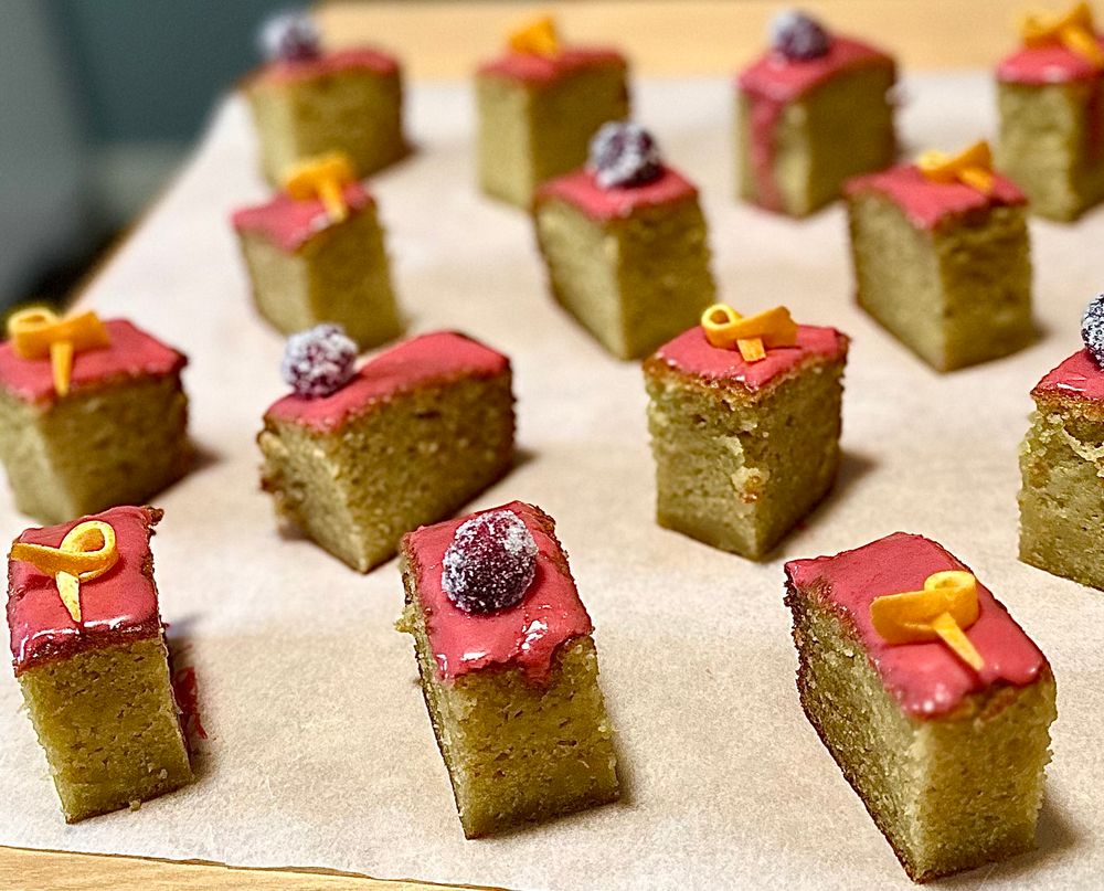 Small rectangular slices of orangey yellow
Cake with bright pink glaze sit on a sheet of parchment paper. They are topped alternately with a sugared cranberry or a small sliver of orange peel.