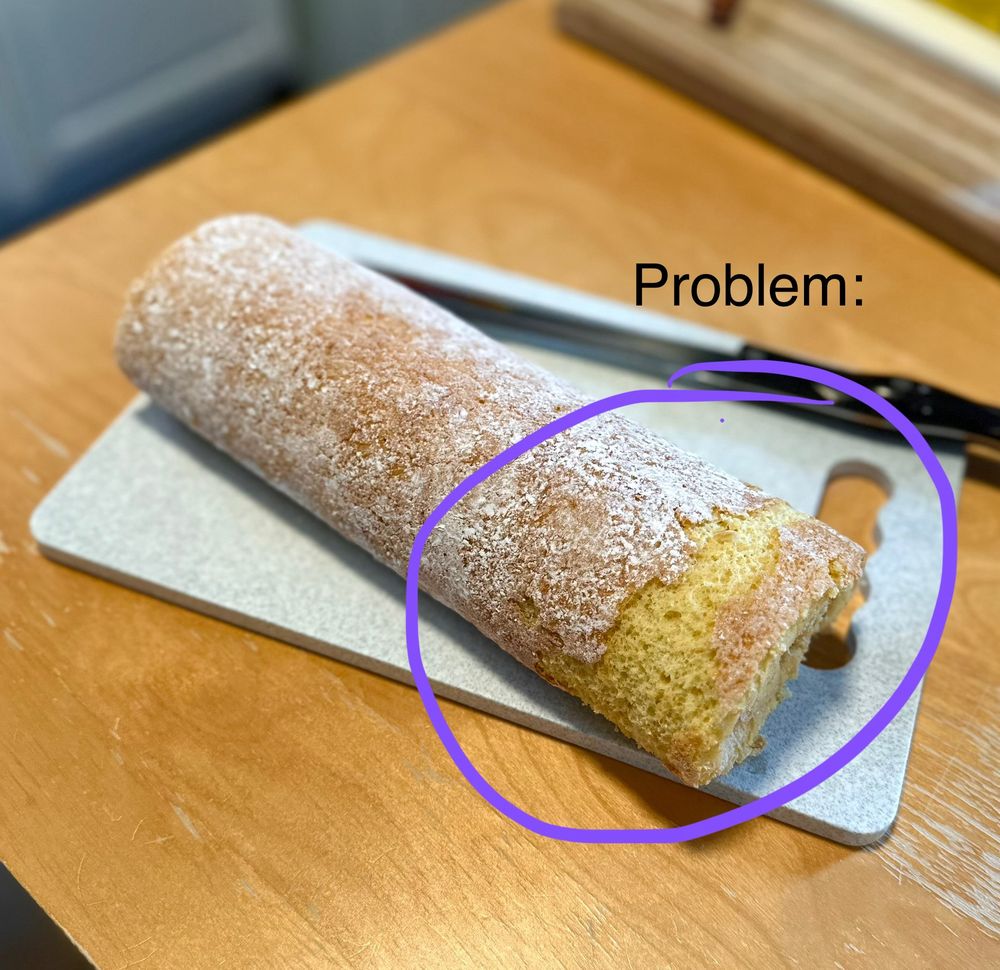 A cake roll sits on a cutting board. A section of the roll’s crust has torn off while being unrolled, and that section has been circled and the text “problem” has been added above it.